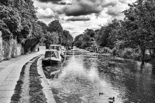 Black and white landscape photograph of a narrow canal lined with trees, featuring several houseboats moored along the left side and a paved walkway beside them.